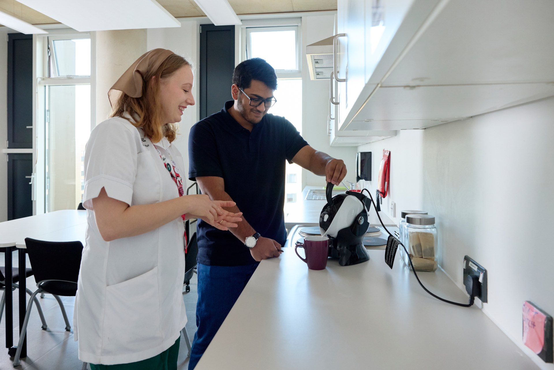Two people stand in a bright kitchen; one is pouring hot water from an electric kettle into a mug while the other watches and smiles.