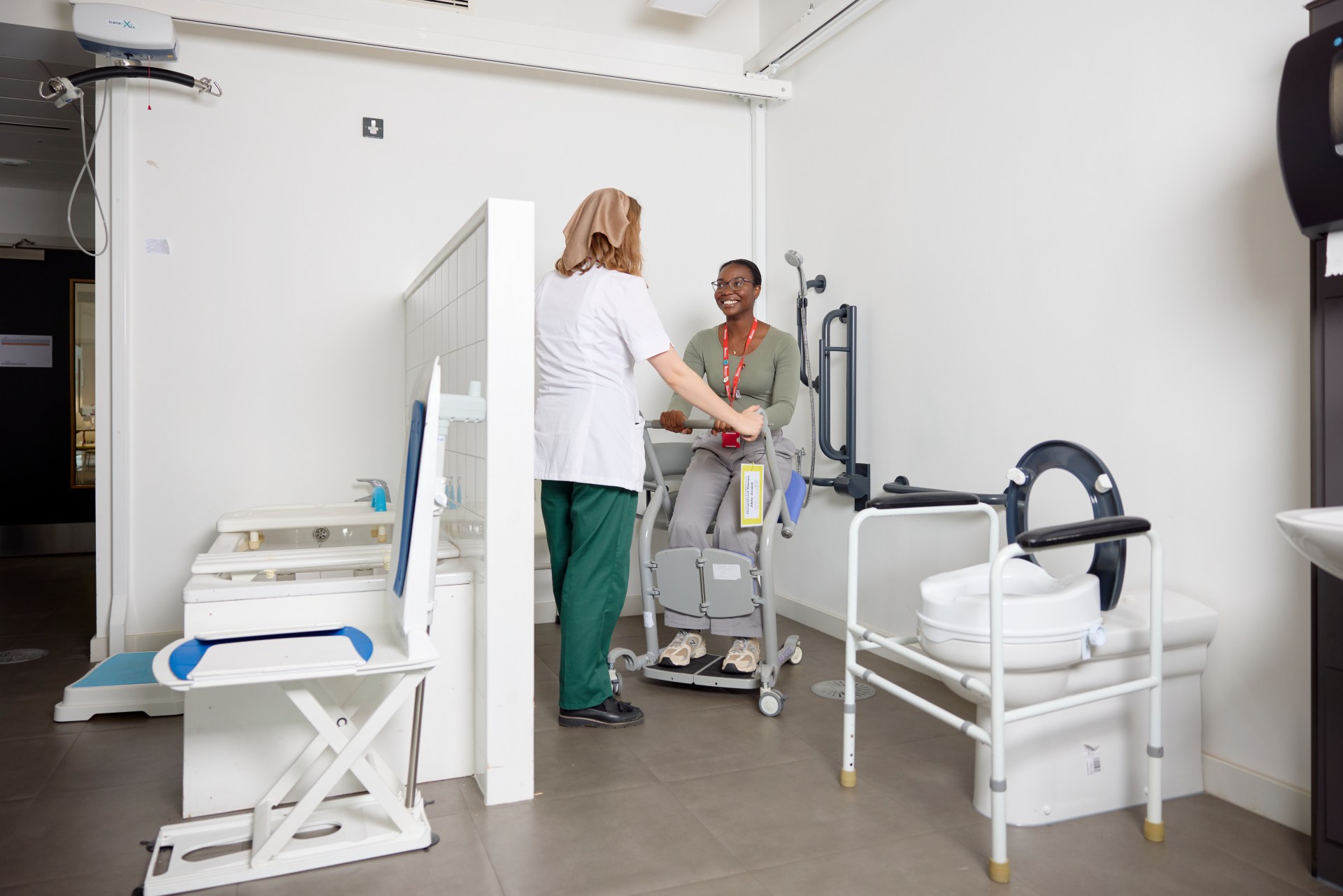A healthcare worker assists a person using a mobility aid in a clinical room equipped with accessible facilities and medical equipment.