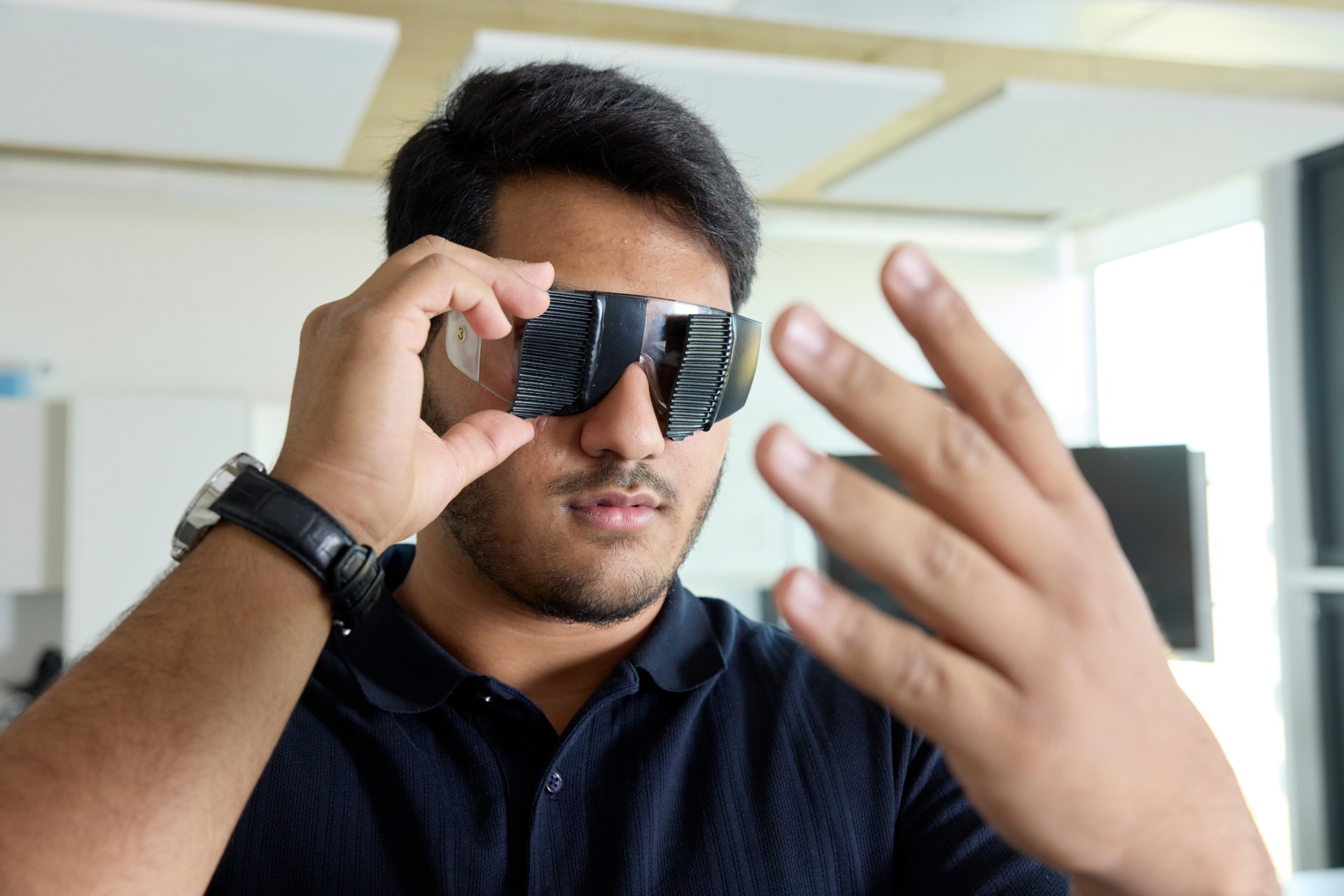 A man in a black shirt wears futuristic, reflective glasses and raises his hand in front of his face, standing in a modern indoor setting.