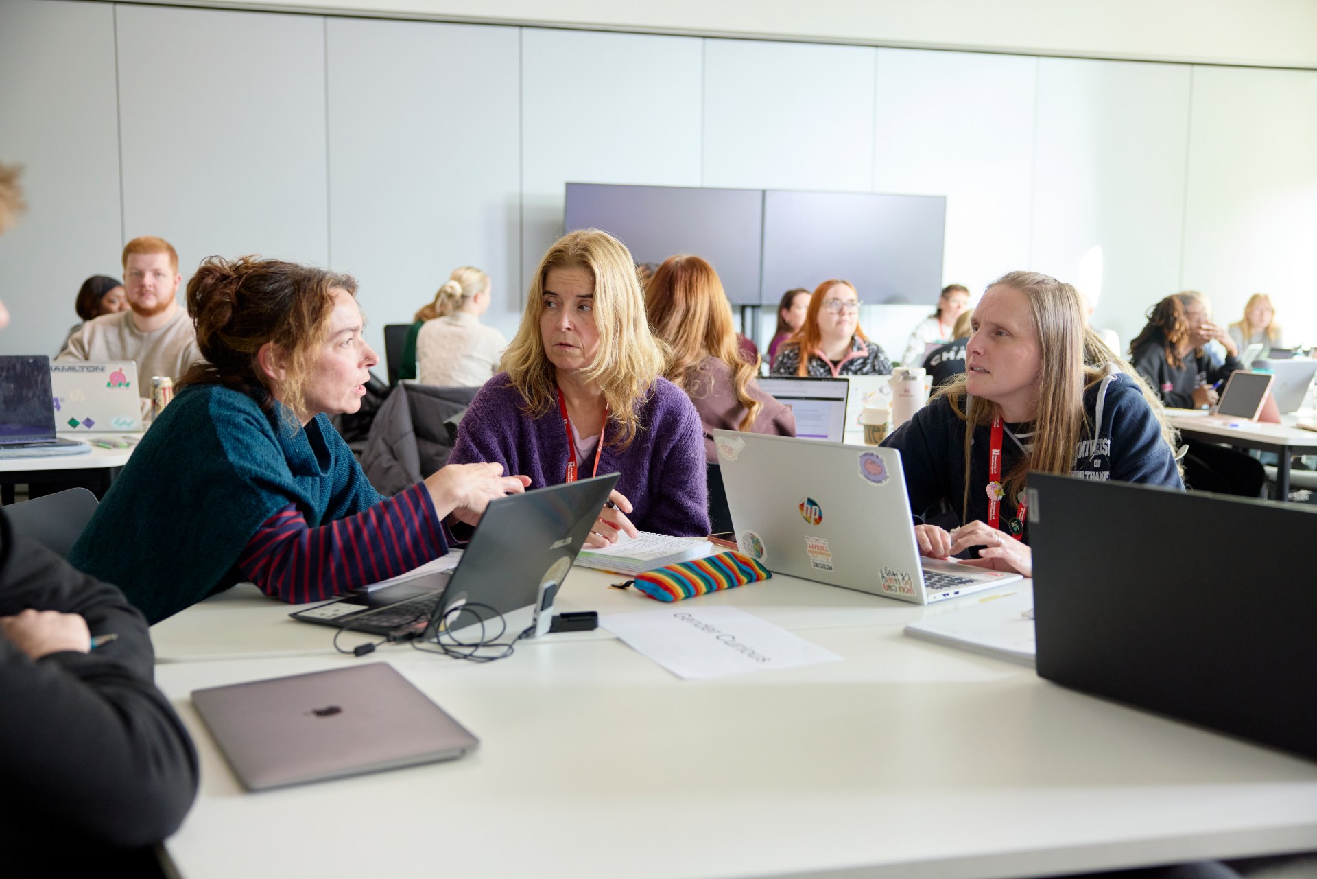 Three people sit at a table with laptops having a discussion in a classroom setting, while others work in groups in the background.