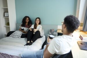 Two students sat on a bed whilst another sits on a chair with a cup of tea in hand chatting in student halls.