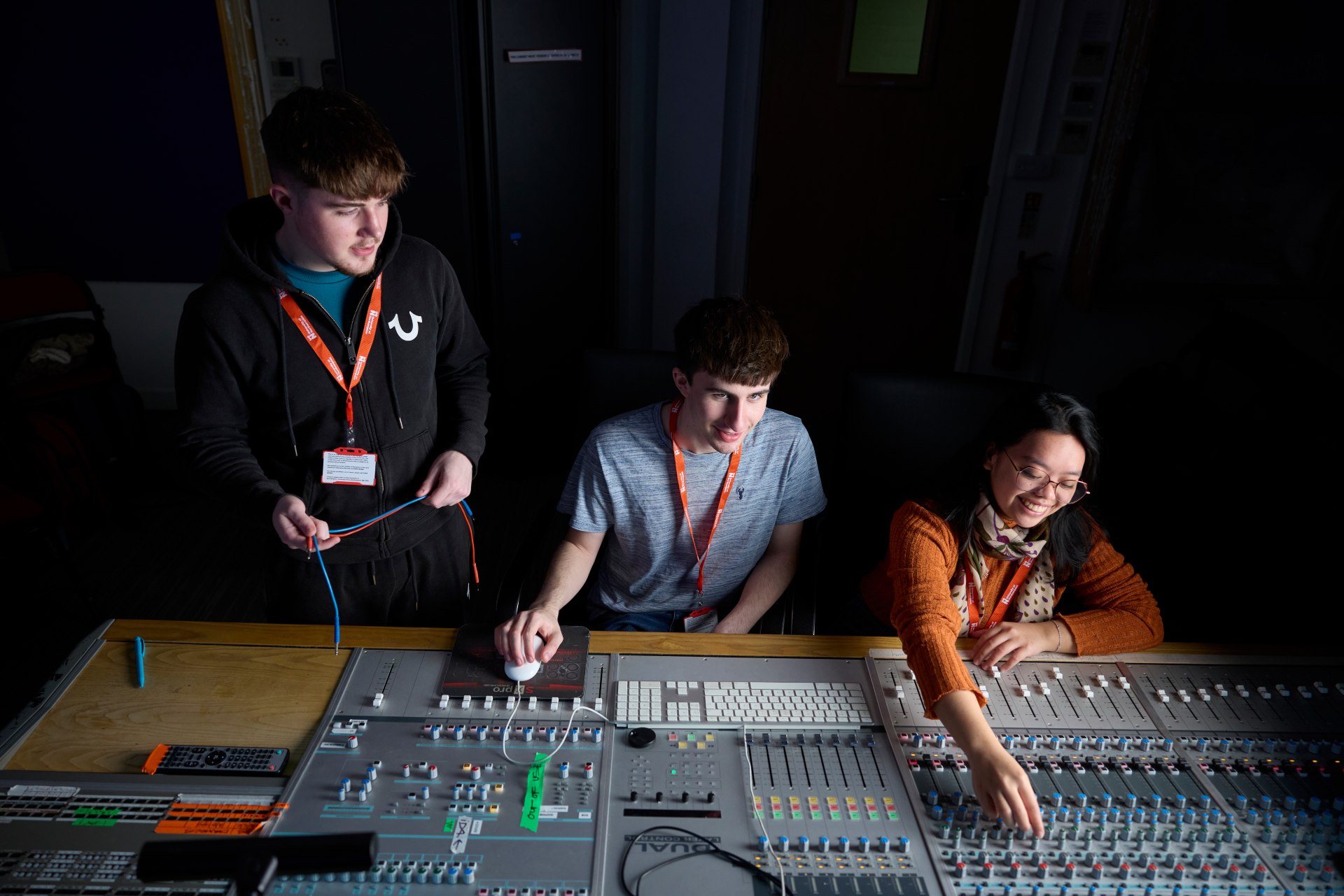 Three students working at a large audio mixing console in a recording studio, adjusting controls and handling cables