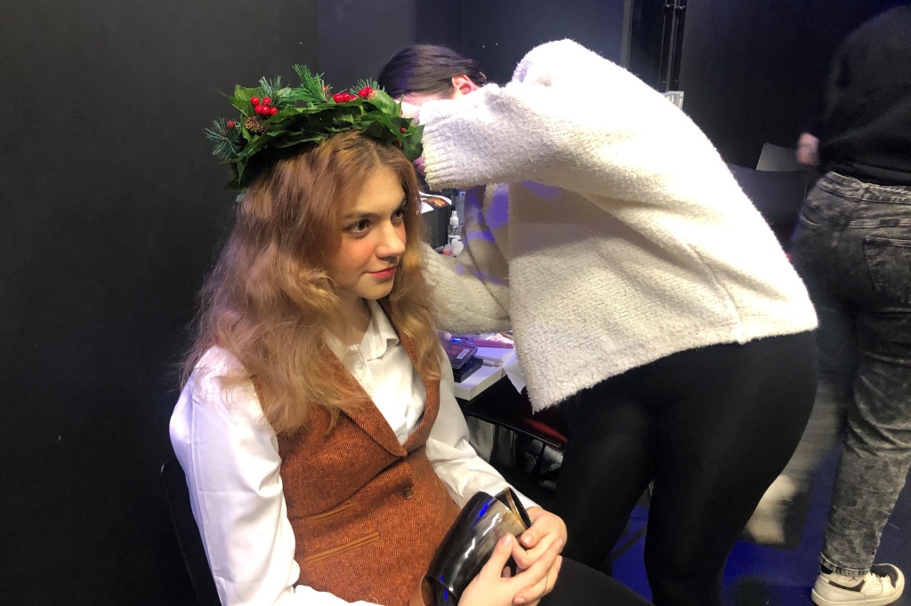A person wearing a laurel wreath and brown vest sits while another person in a white sweater styles their hair.