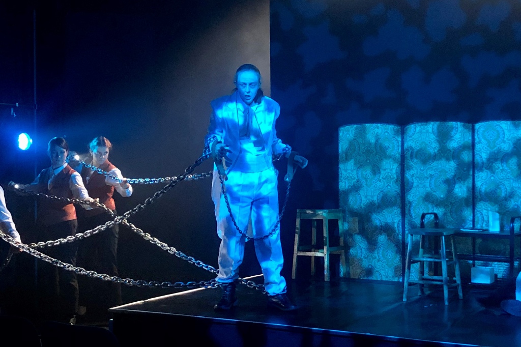 A person dressed in white stands on stage wrapped in chains, with blue lighting and two people in the background holding the chains.