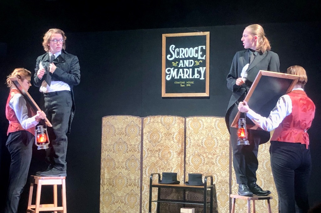 Four people in stage costumes set up a "Scrooge and Marley" sign, with two holding lanterns and standing on stools, on a theater set with a gold patterned divider in the background.