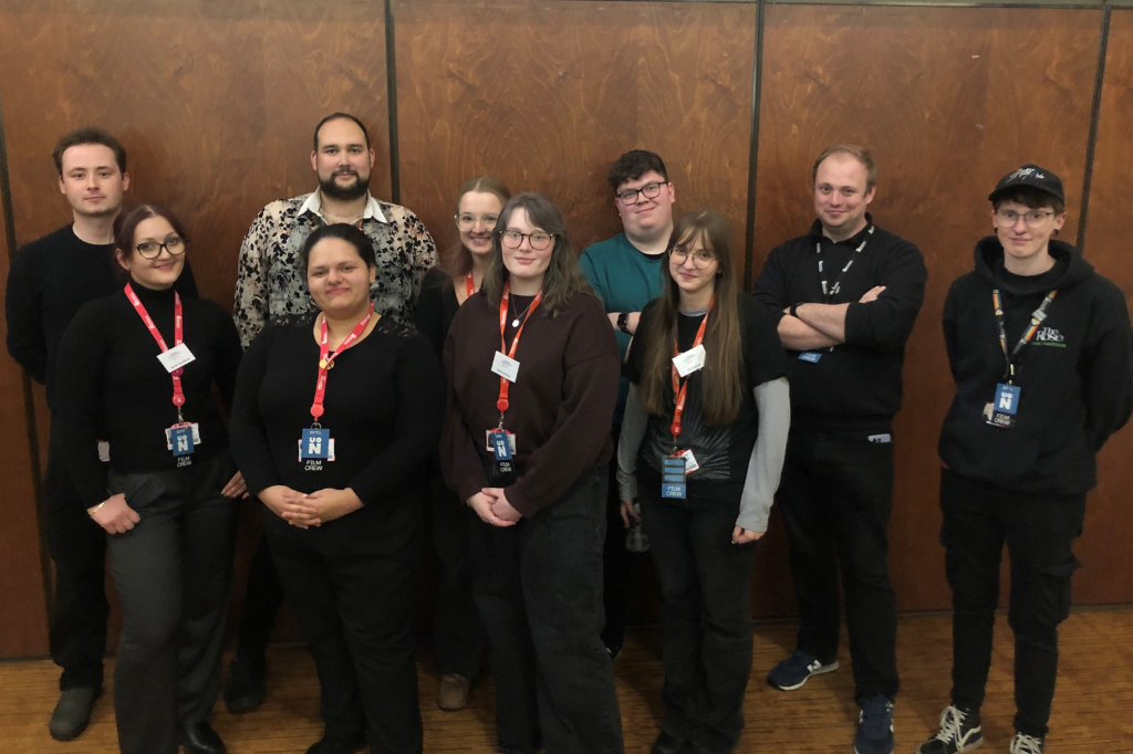 Eleven people stand indoors in two rows, facing the camera. Most are wearing black outfits and staff lanyards. The background is a wooden wall.