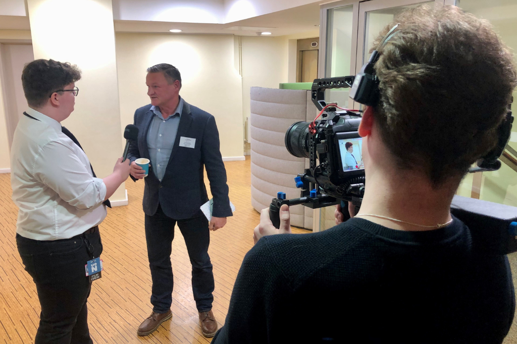 A person holds a microphone and interviews another person holding a cup, while a third person films the scene with a camera in an indoor setting.