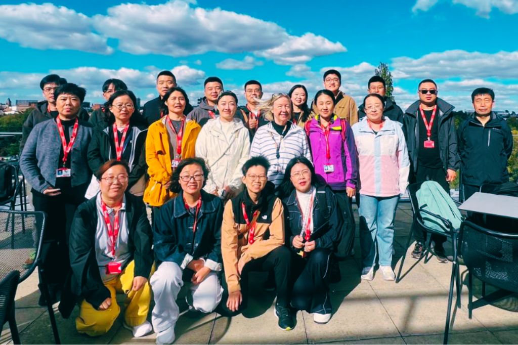 A group of Chinese academics standing shoulder to shoulder with UK counterparts on a roof-top balcony with fluffy white clouds in the sky.
