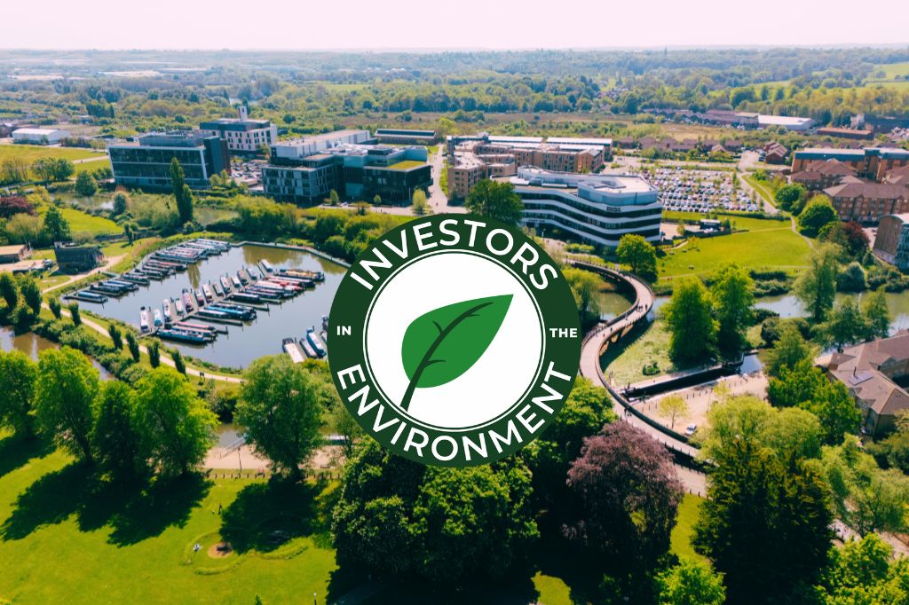 Aerial view of office buildings, marina, and greenery with the "Investors in the Environment" logo featuring a green leaf overlaid in the center.