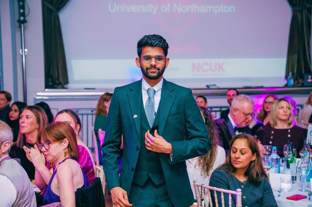 A man in a suit stands in front of a seated audience at an event, with a projector screen displaying "University of Northampton" and "NCUK" in the background.