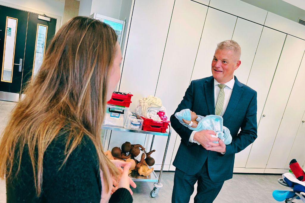 Stuart Andrew MP holds a baby mannequin while talking to a woman in a room with medical training equipment and supplies on a cart.