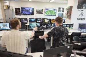 Two people sit in a computer lab with multiple monitors displaying video games and digital art, discussing their screens.
