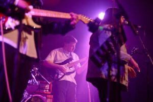 A band performs on stage under purple lighting, with musicians playing electric guitars and a person singing into a microphone.