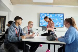 A Marketing student sits on a table to talk to a group of students. She is holding a VR headset. The other three students have open laptops in front of them and one has a VR headset.