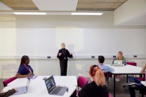 An academic dressed in black with a white turban lectures in a classroom in front of a white background. In front of her, students sit at tables, taking notes on laptops and tablets.