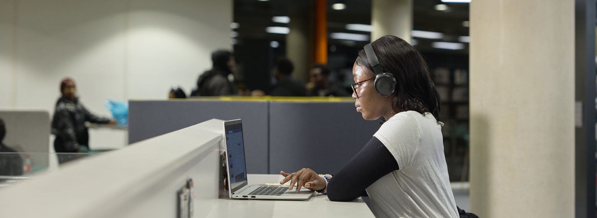 A female student wearing a pair of black headphones works on a laptop in the library.