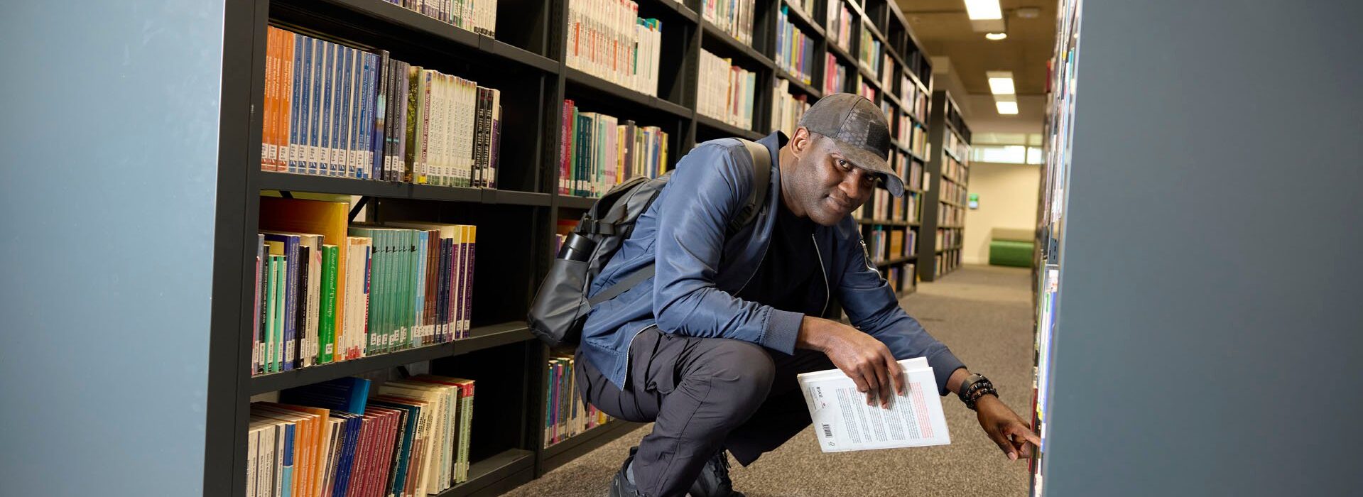 A student crouches by a bookstack to select a book. He is waering a baseball cap and backpack and is looking at the camera.