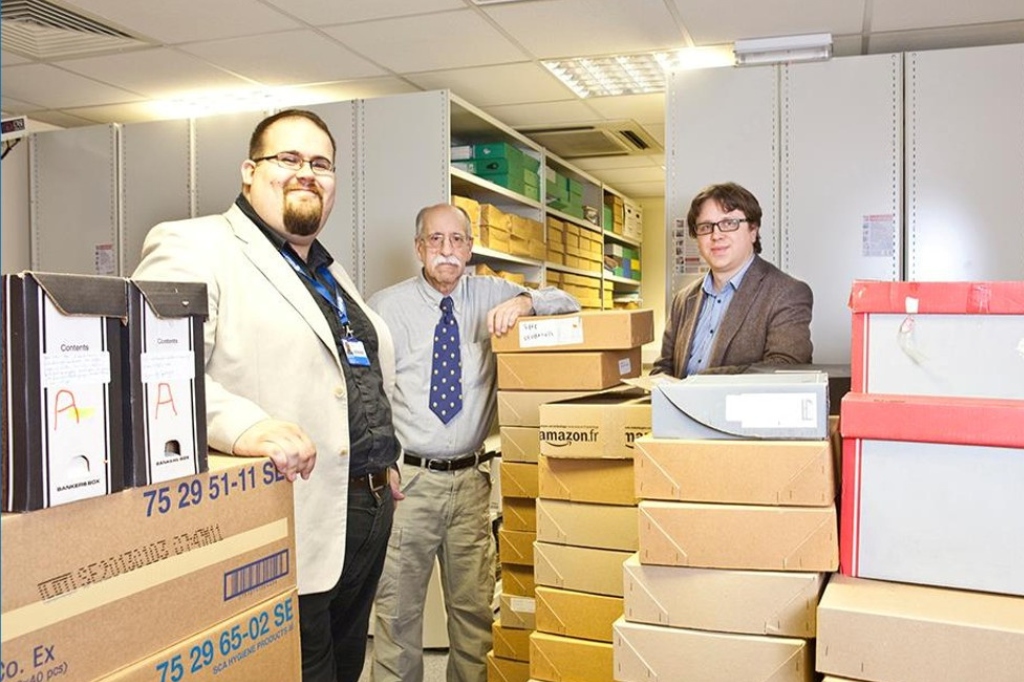 Three people stand in an archive surrounded by cardboard boxes, smiling at the camera.