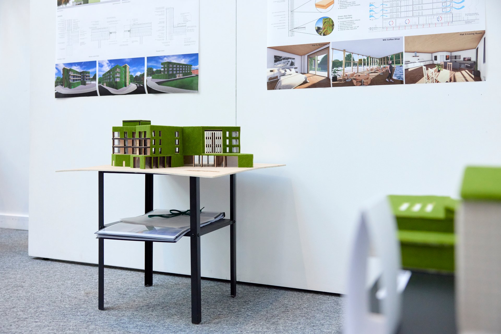 Student architectural model of a green-coloured building displayed on a table beneath design drawings and digital renderings.