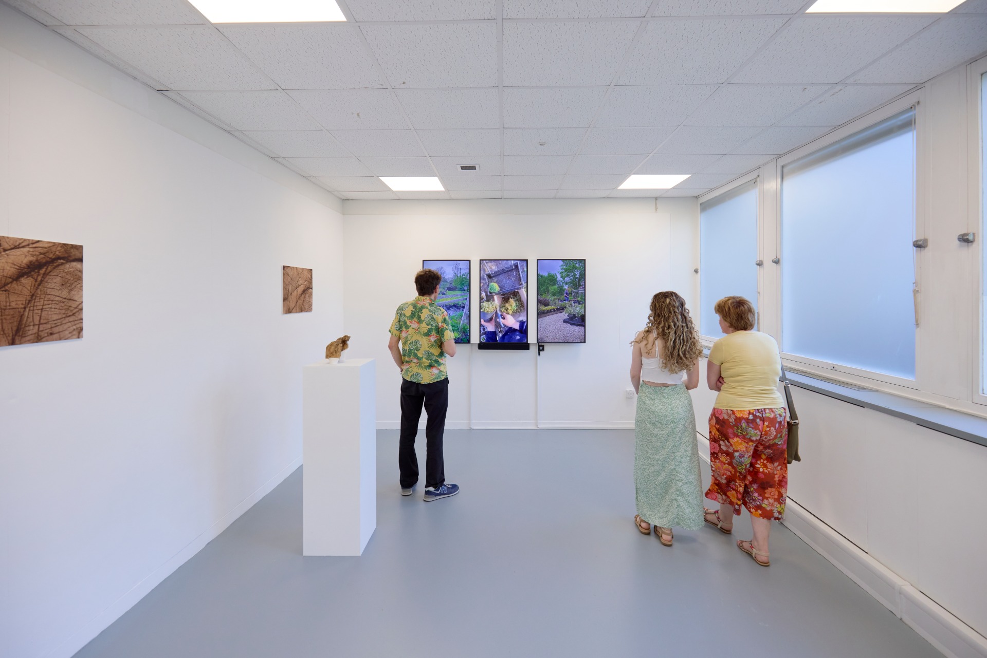 Visitors stand in a gallery viewing three vertical video screens and a small student sculpture on a white plinth.