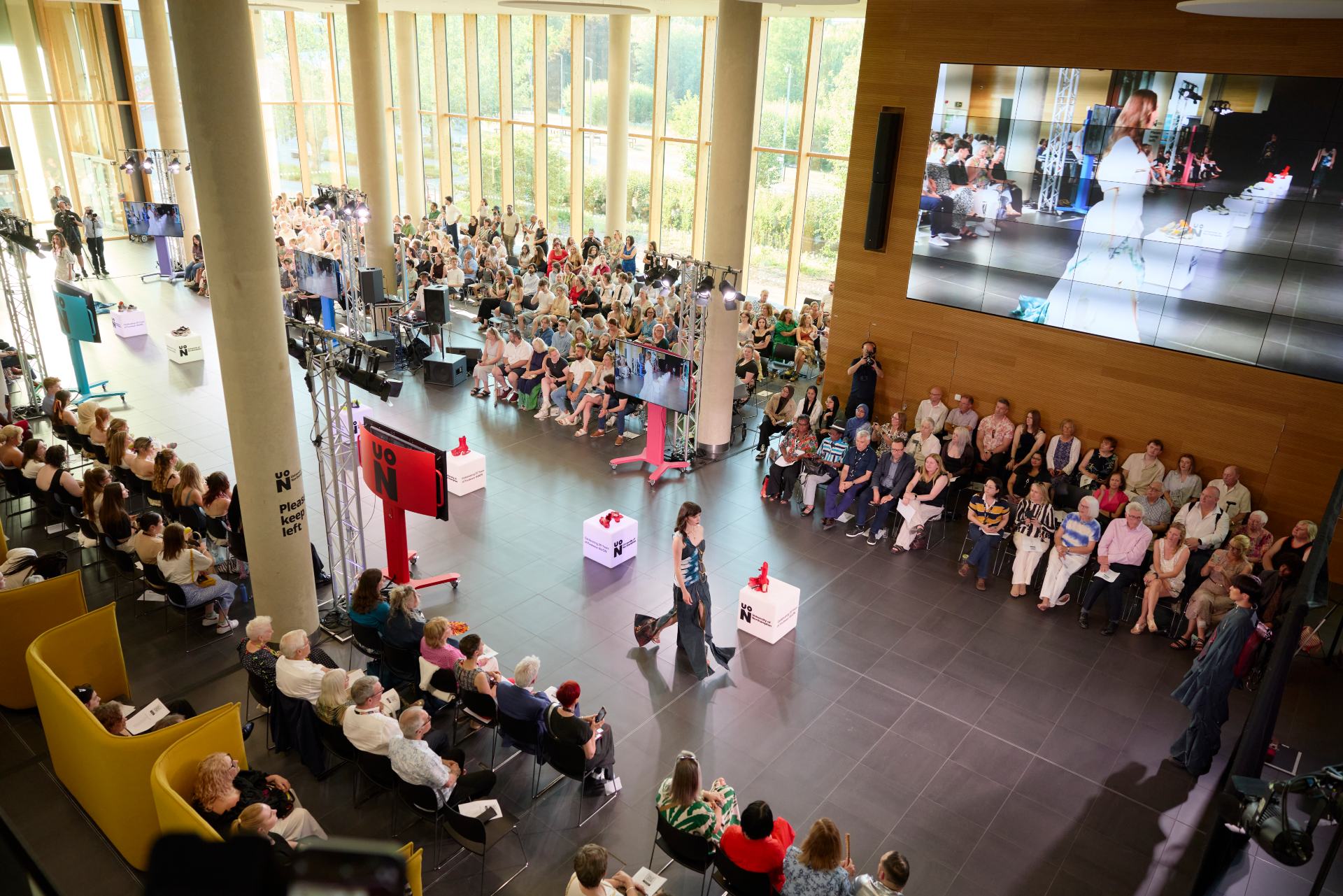 Wide view of a student fashion show in a large atrium, with a catwalk running through seated audiences on both sides.
