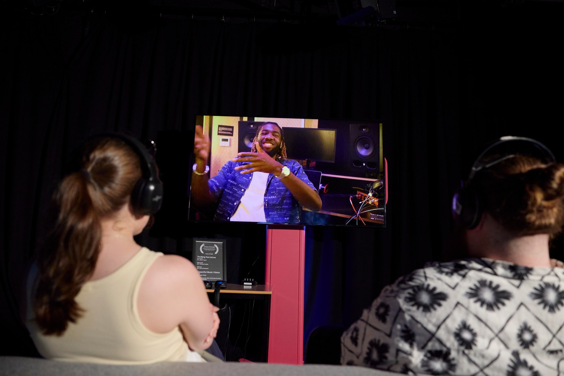Two visitors wearing headphones watch a student video artwork displayed on a screen in a darkened exhibition space.