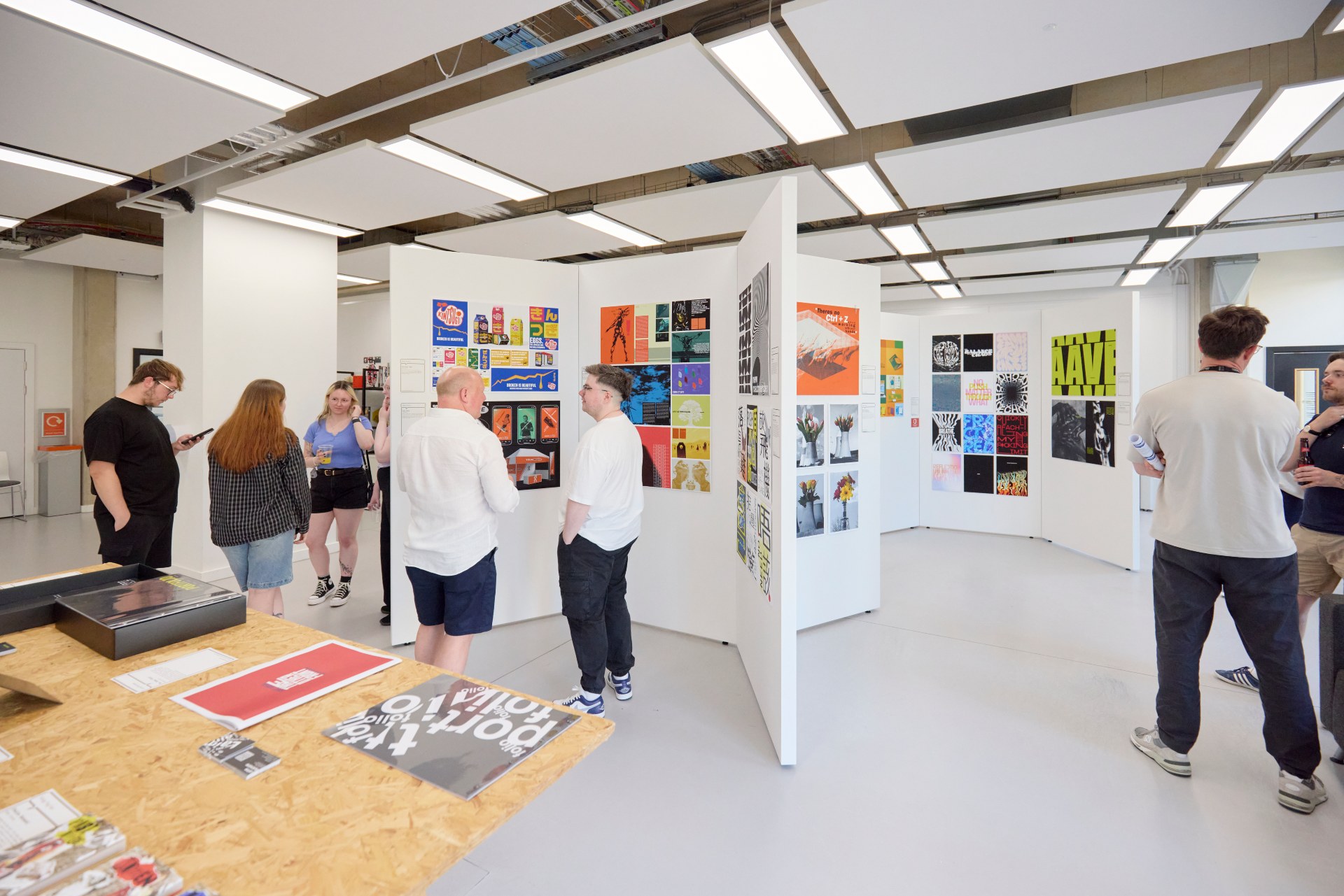 Visitors stand in a gallery surrounded by colourful student graphic design posters and printed artworks displayed on white panels.