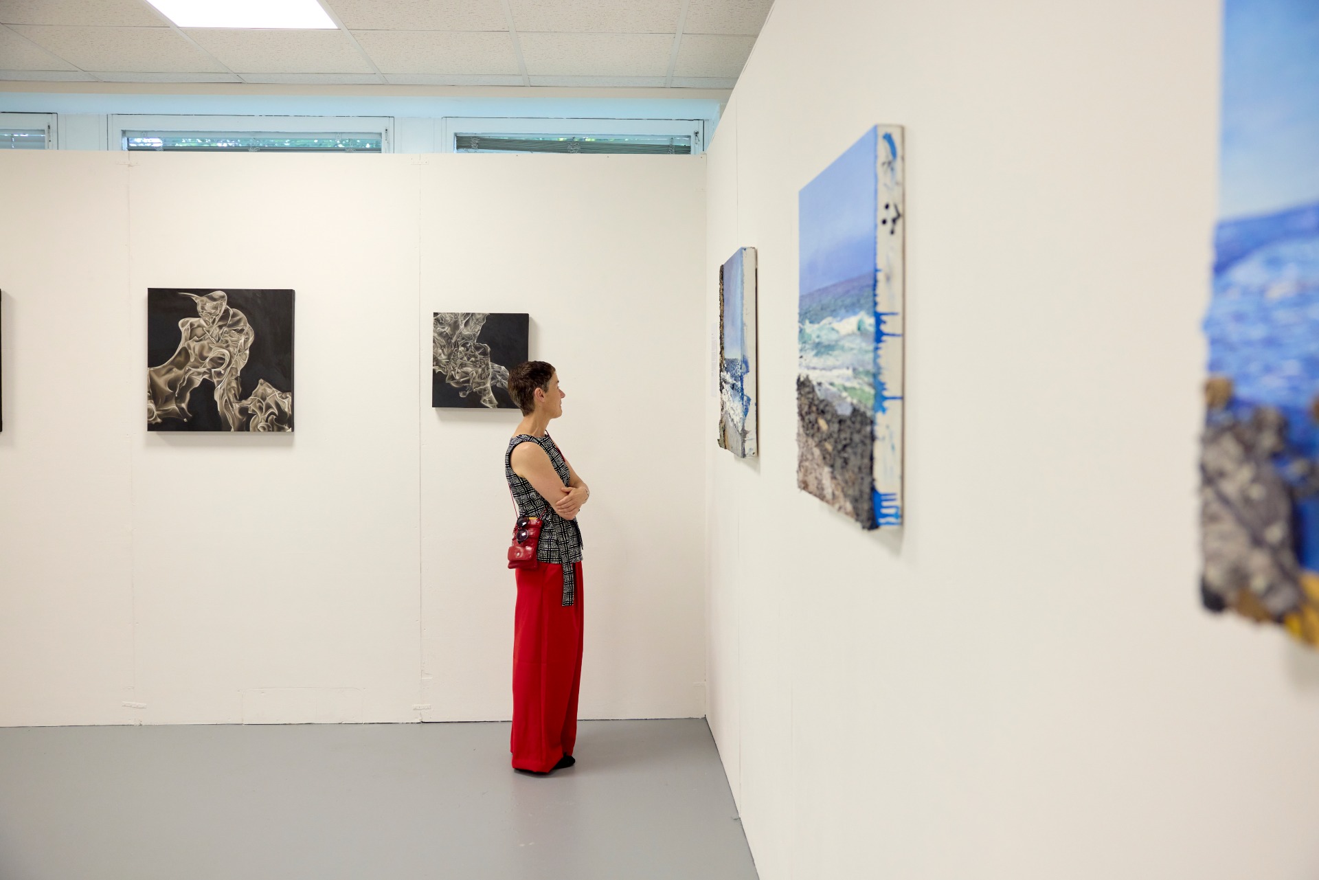Visitor stands in a white gallery space viewing student artwork displayed on the walls, including abstract monochrome pieces and mixed-media coastal scenes.