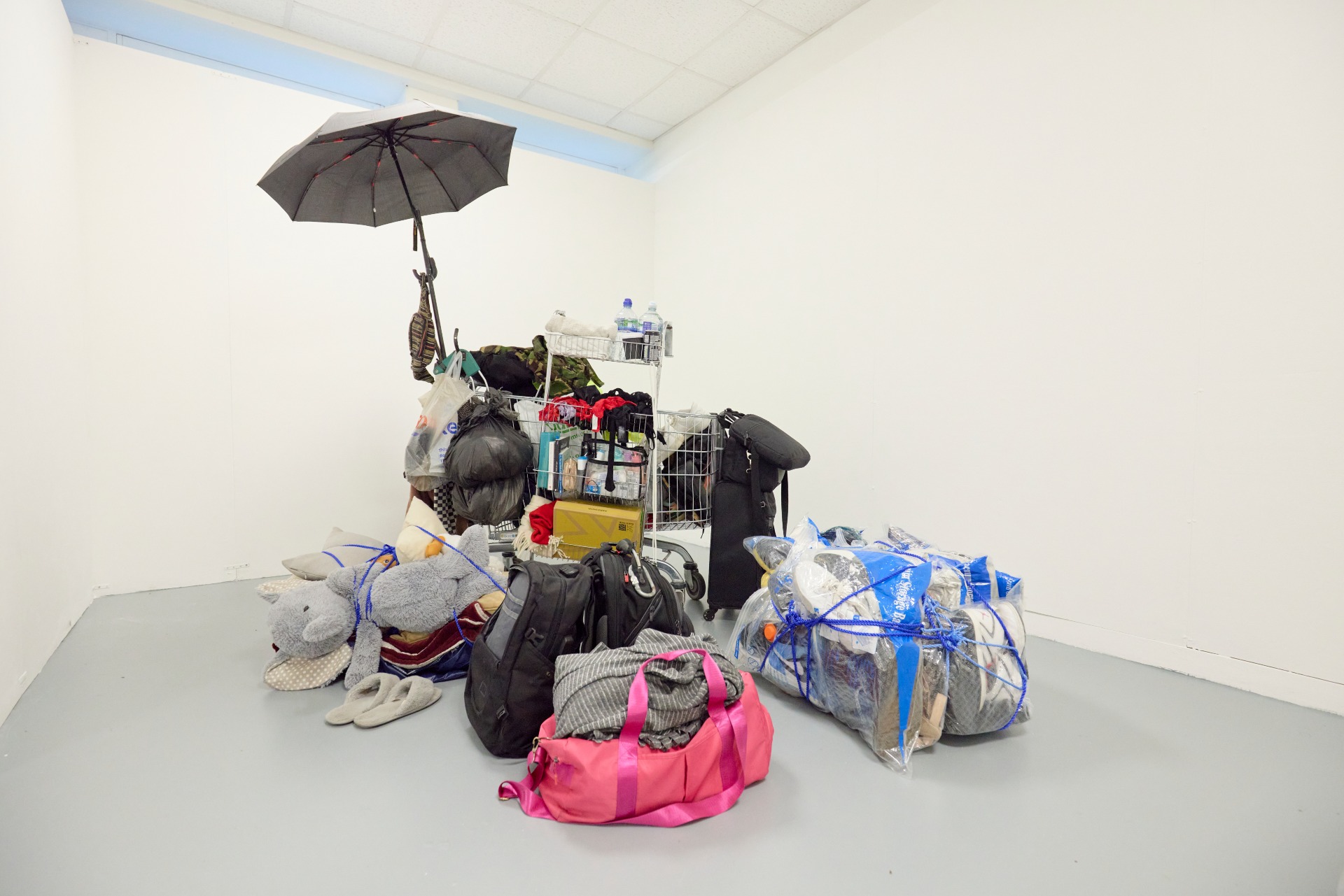 Student installation made from bags, clothing, soft toys and household items arranged in a gallery corner, with an umbrella fixed above.