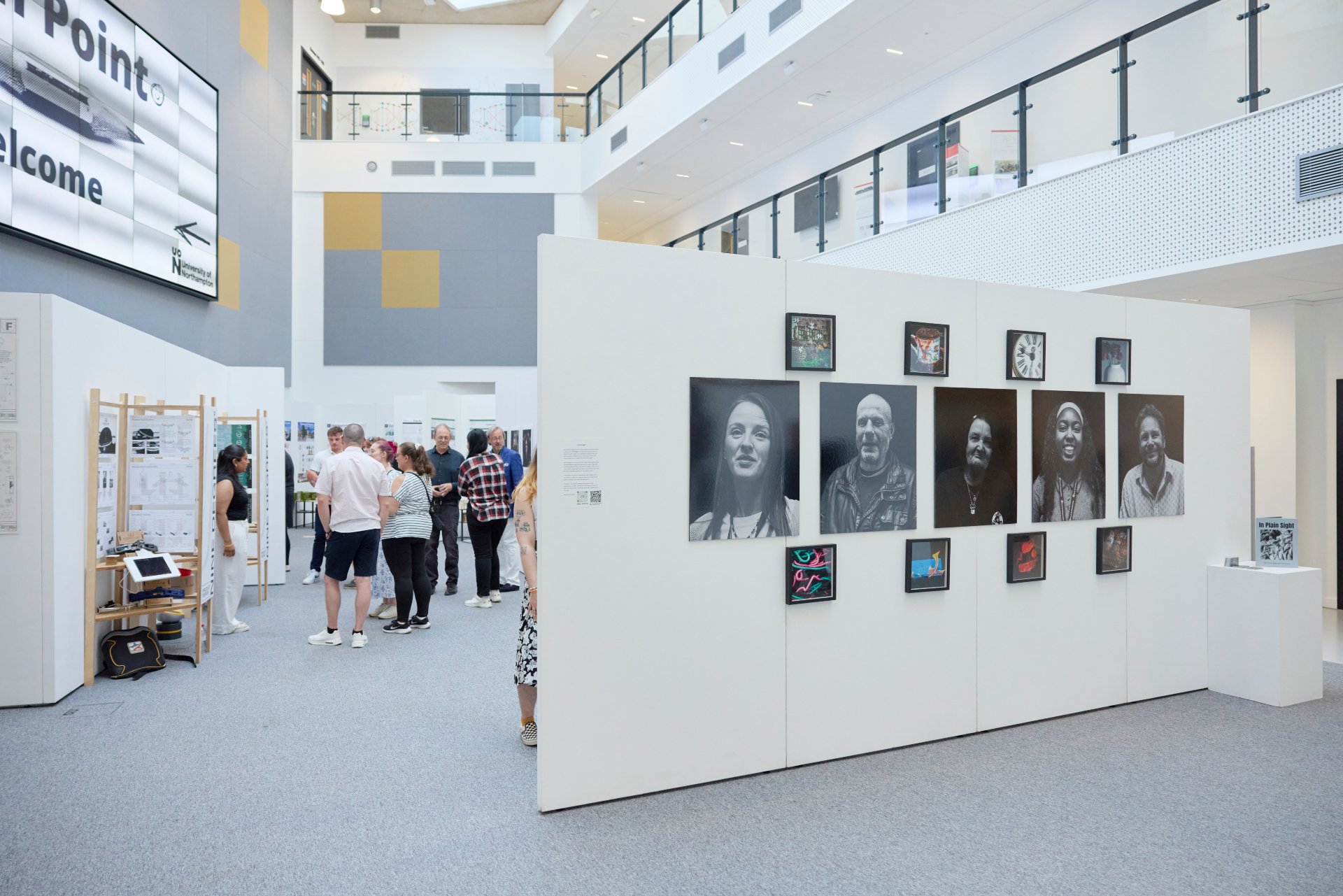 Visitors walk through a gallery space with student portrait photographs and mixed-media artworks displayed on white panels.