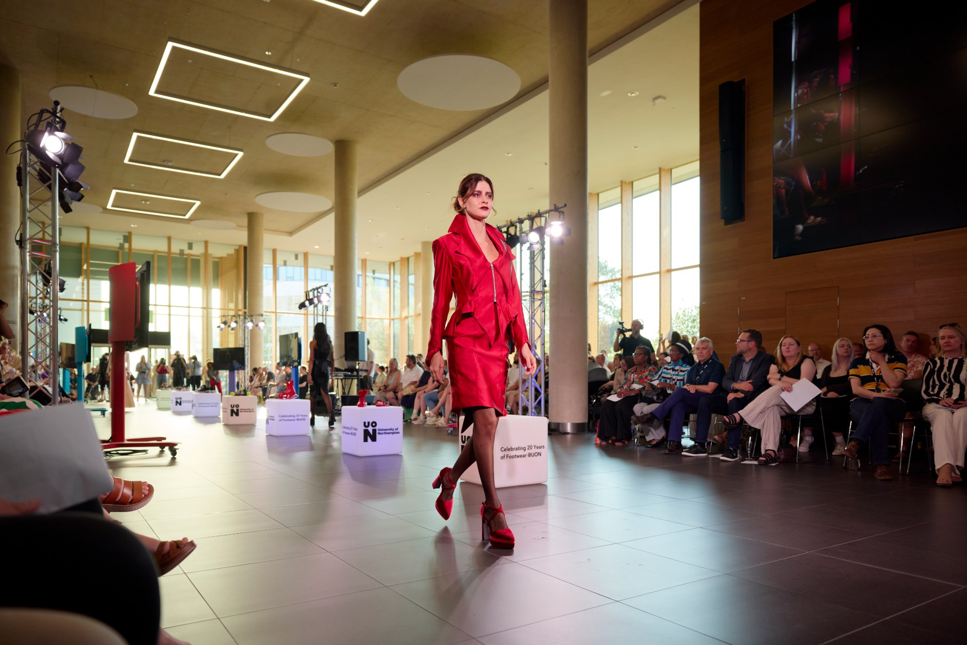 Student fashion model walks down a catwalk wearing a red tailored outfit and heels, with audience seated along the runway.
