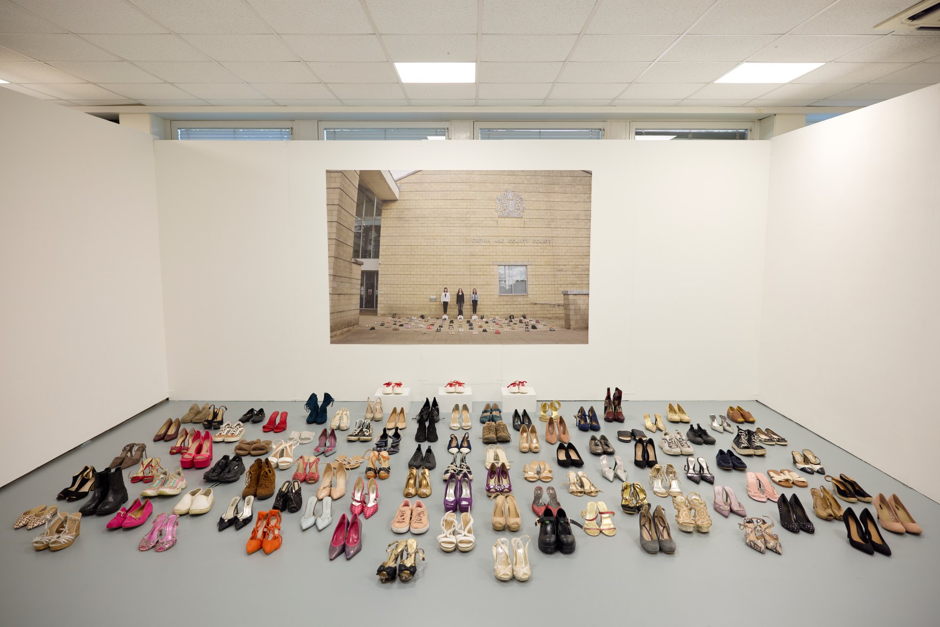 Rows of different shoes are arranged on the gallery floor beneath a large student photograph mounted on the wall.