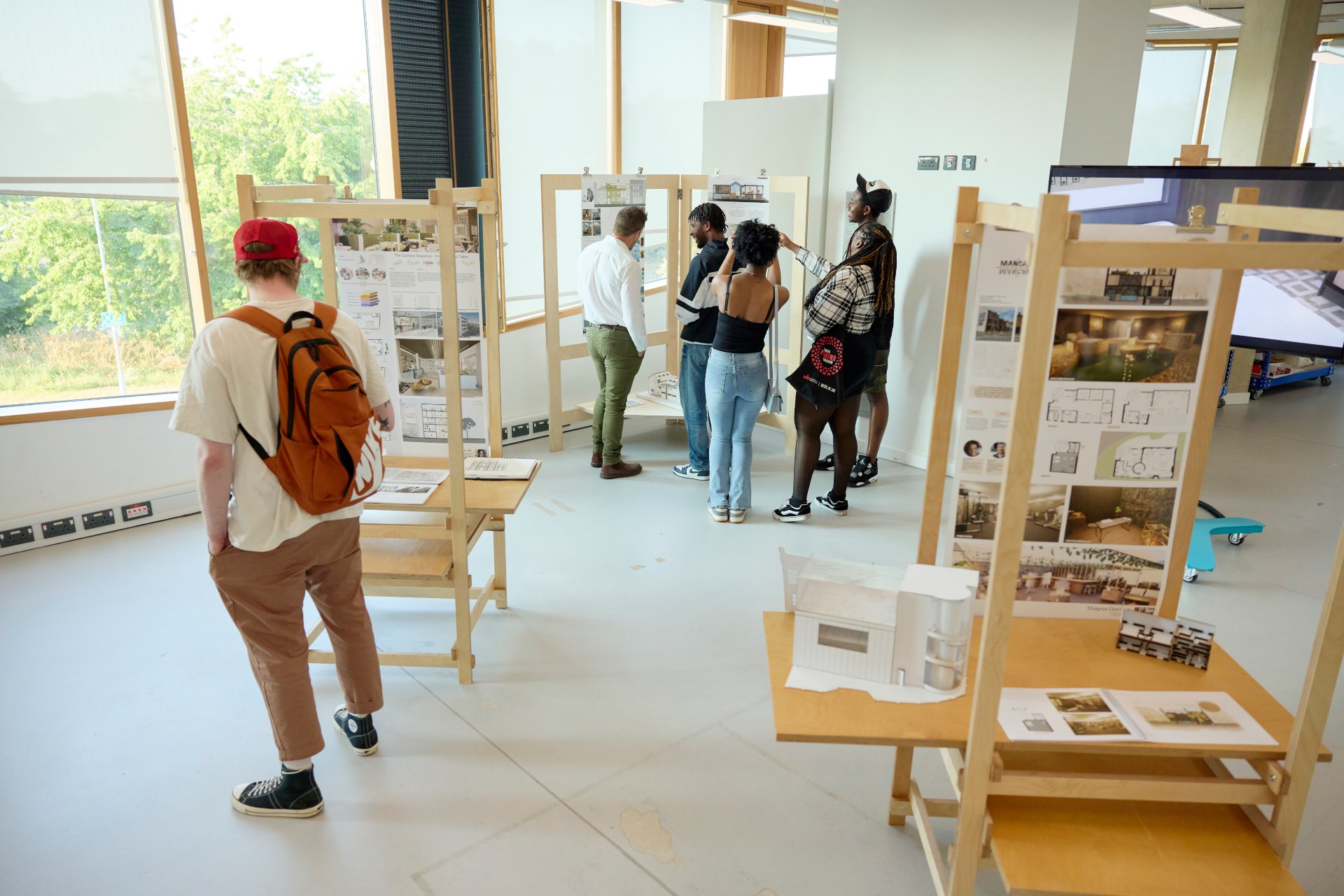 Visitors view student architecture projects displayed on wooden stands, with drawings, models and digital screens in a studio gallery.