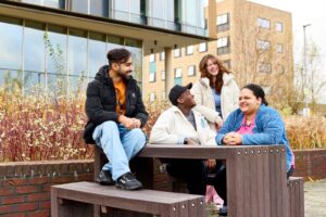 Four students sit on or stand around a table outside the Learning Hub on Waterside campus. They are talking and laughing.