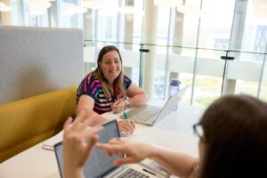 A postgraduate student in a striped t-shirt smiling and speaking to another student sat opposite