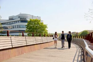 Three students walk on a curved, wooden footbridge on a bright, sunny day. Visible in the background is the Creative Hub and several trees.