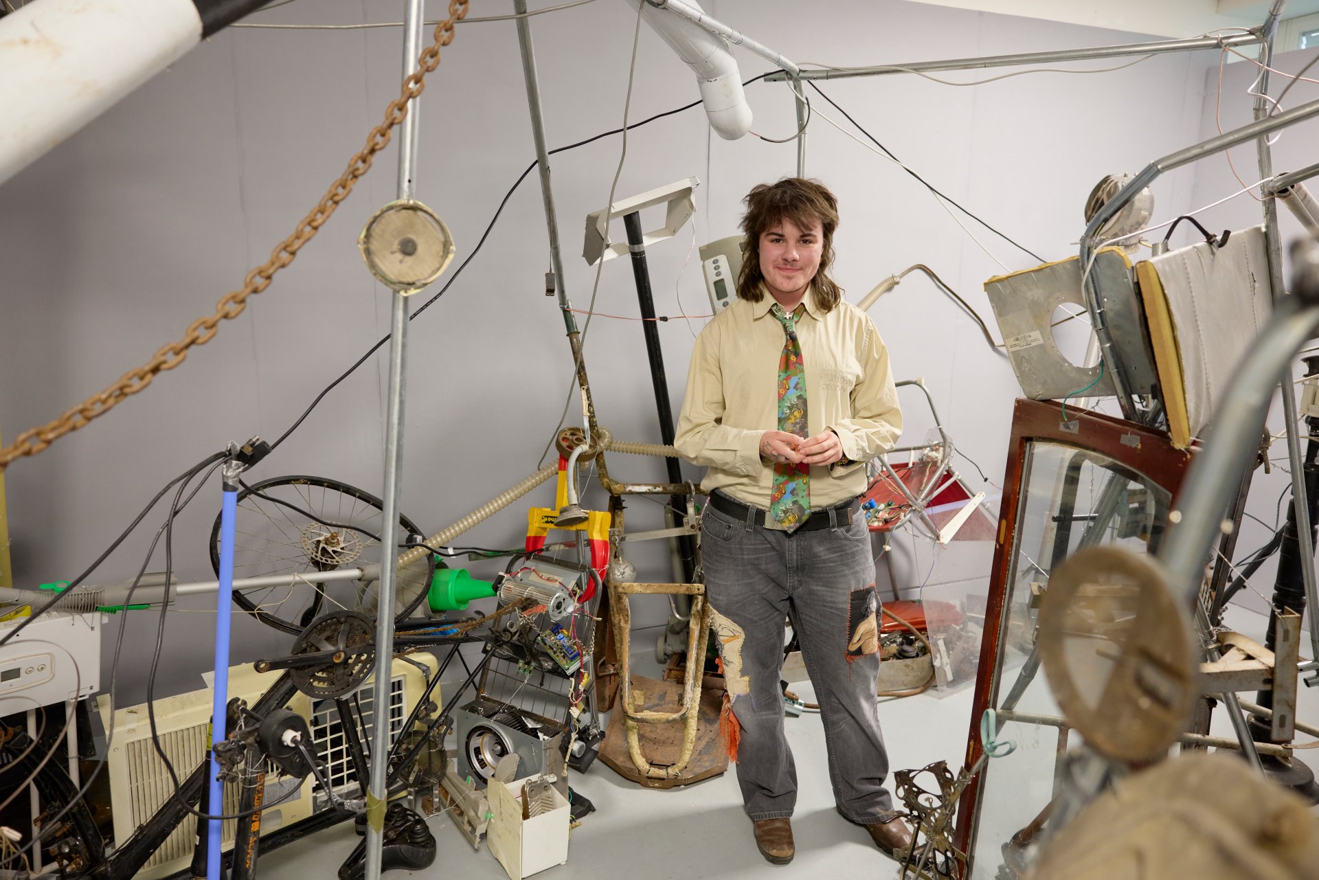 Fine Art student stands inside an installation made from found objects, metal frames, wires and mechanical parts arranged throughout the gallery space.