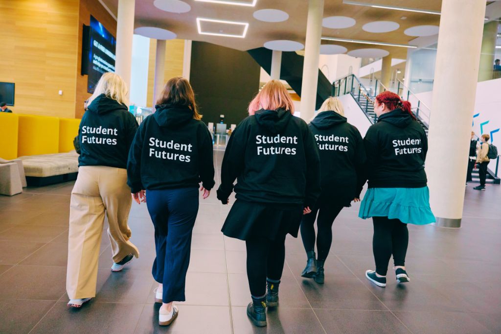 A group of students wearing hoodies with Student Futures written on their backs walk away from the camera in unison.