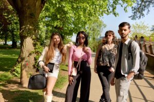 Four students stand on a path in Beckets Park next to the curved footbridge. It is a bright and sunny day. The stduents are dressed casually and are carrying bags.
