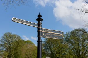 View of a Northampton signpost against a blue sky with clouds and trees. The signpost is black with gold writing, and has arms which point to the town centre, the Skatepark, and Waterside campus.