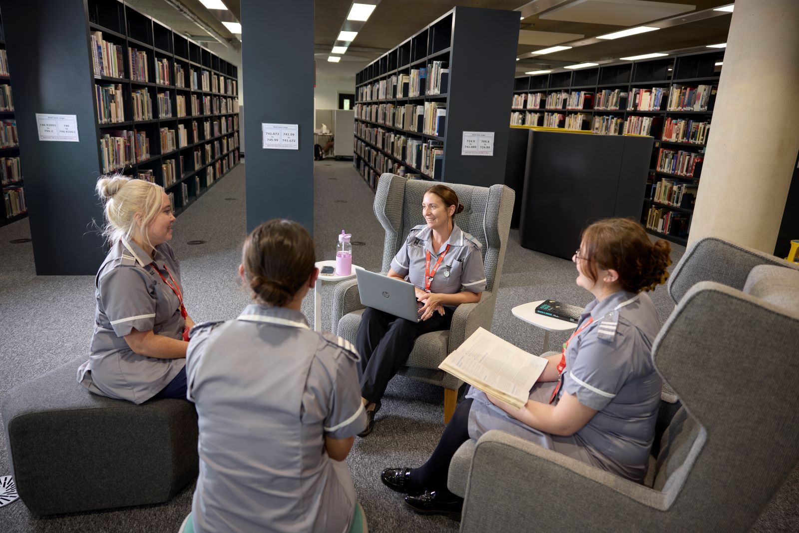 Four student nurses sit on armchairs and pouffe seats in the library in the Learning Hub. They are wearing grey uniforms. One has an open laptop on her lap, and another holds an open book.
