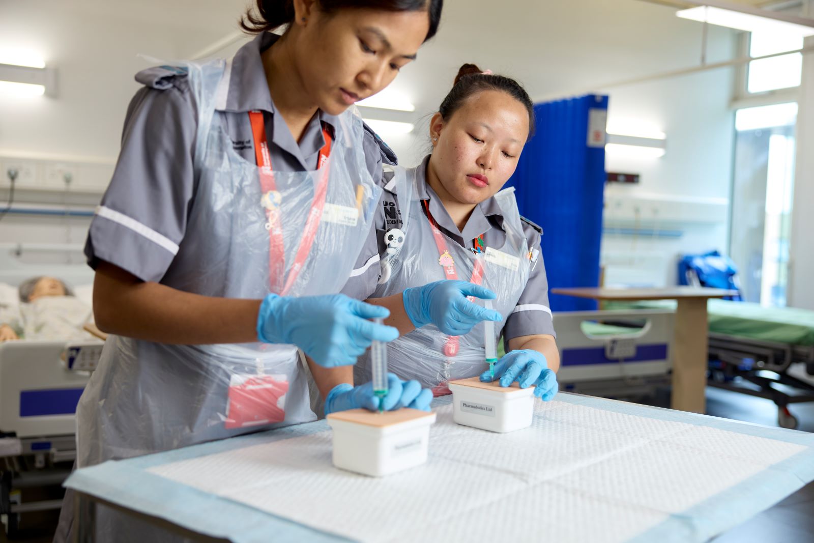 Two student nurses practice using syringes on a ward in the Nursing Suite. They are both wearing grey uniforms, plastic aprons and light blue gloves.