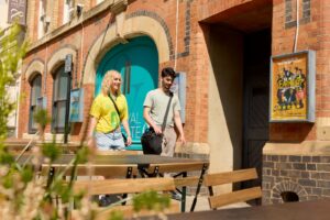 Two students walk past the Royal and Derngate theatre on a sunny day. In the foreground are sets of tables and chairs. Both students are casually dressed in t-shirts and carrying bags.