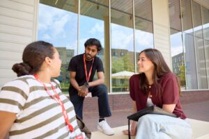 Three students chat outside the Learning Hub at Waterside campus on a sunny day. Two sit on the steps, while the other crouches.
