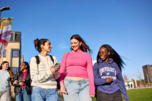 Three students walk on campus on a bright day. One is holding a smartphone and all three are smiling. Behind them, the sky is blue and the Senate building is visible.