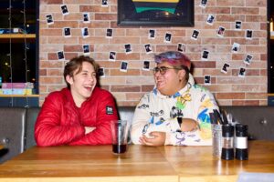 Two students are sitting at a table in the Waterside restaurant. They are chatting and laughing. On the table is a half-empty pint glass, cruet set, and silver bucket containing cutlery and napkins.