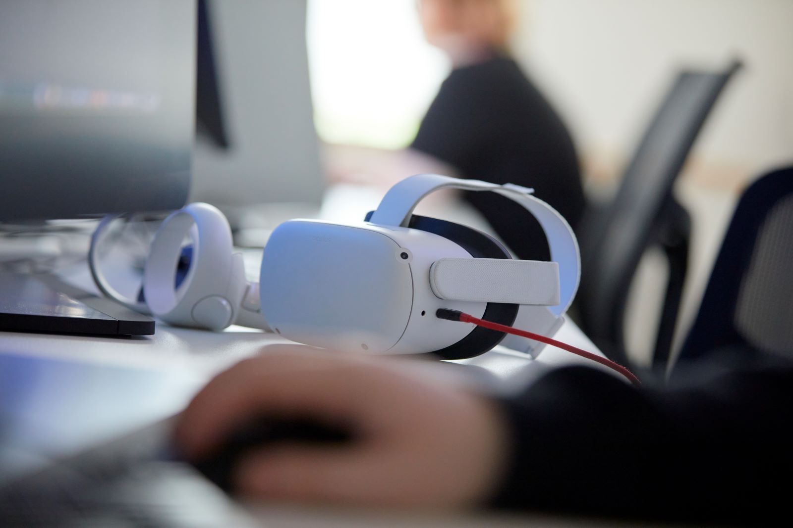 Virtual reality headset and controllers on a desk. On either side two blurred people work at computers.