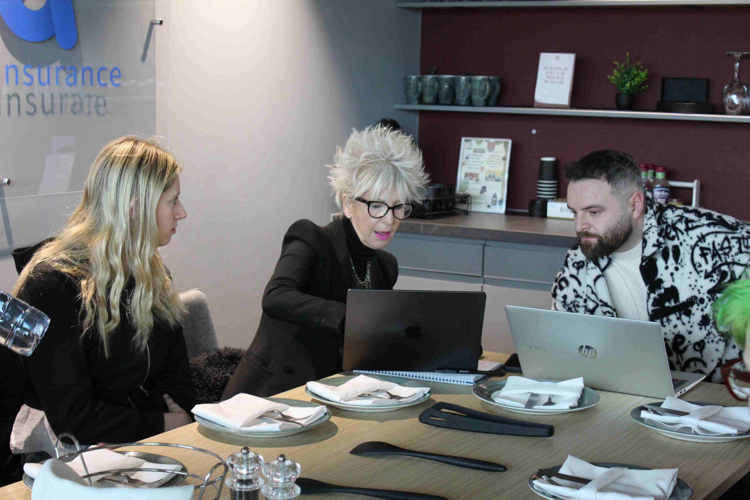 Three people sit around a table and discuss over a laptop.