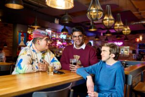 Three students sat round a table in a student bar with drinks in hand and on the wooden table in front of them chatting to one another.