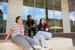 Three students relax on steps outside the Learning Hub at Waterside campus on a sunny day. Two sit on the steps, while the other crouches. They are looking into the distance off-camera and smiling.